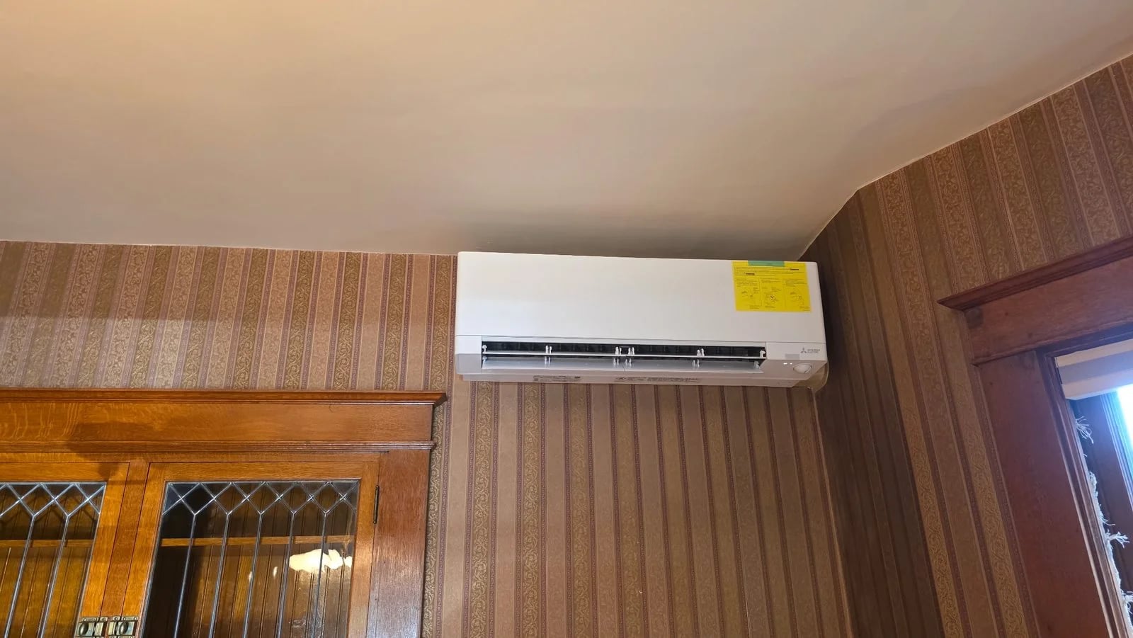 A ductless indoor unit high on a wallpapered wall above wood paneling and a leaded-glass cabinet