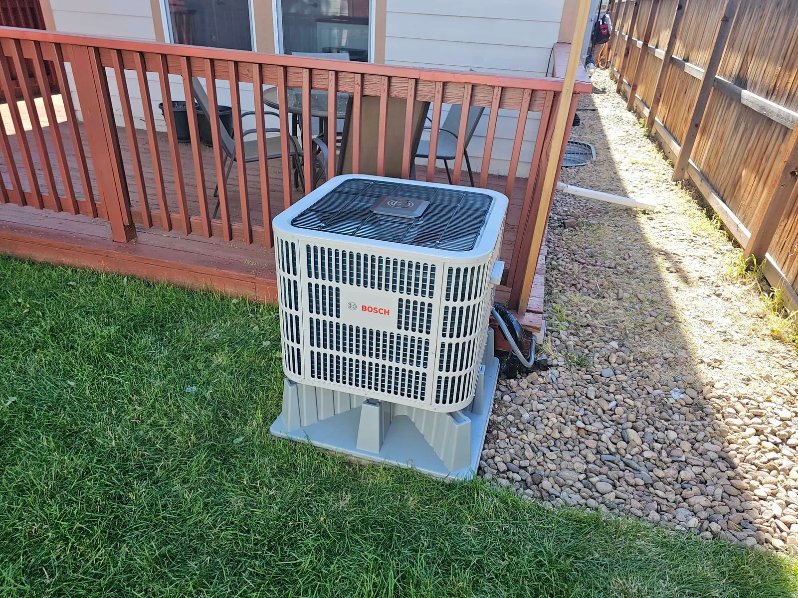 A Bosch outdoor unit on a gray riser on a gravel strip between a green lawn and a red-stained deck railing