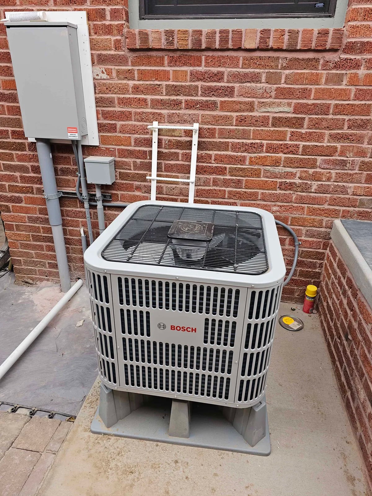 Bosch outdoor unit on a gray riser on a concrete pad against a brick wall, with a disconnect box and conduit