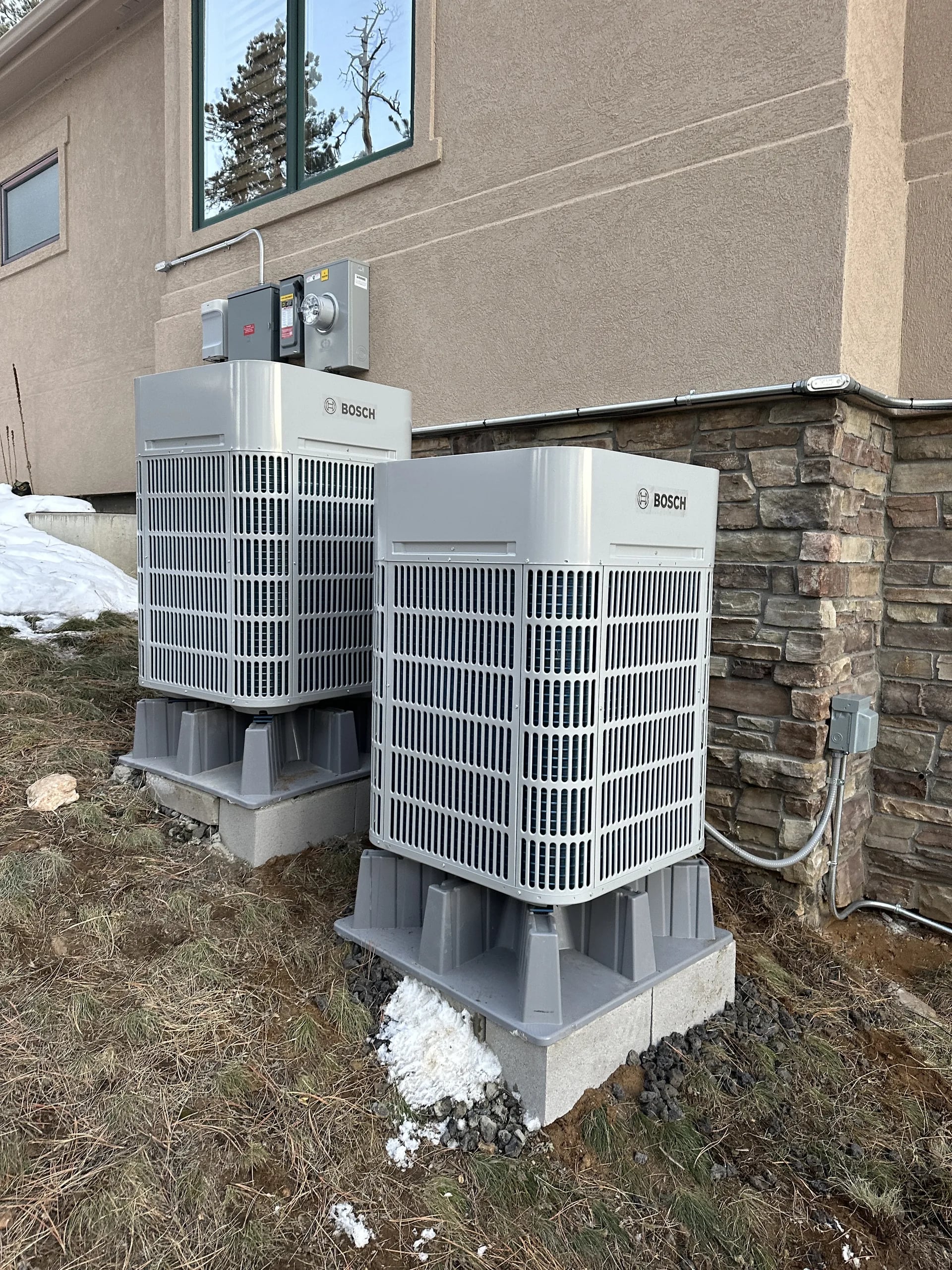 Two Bosch outdoor units on gray risers set on concrete blocks beside a stone-veneer wall, with electrical boxes and conduit on the wall
