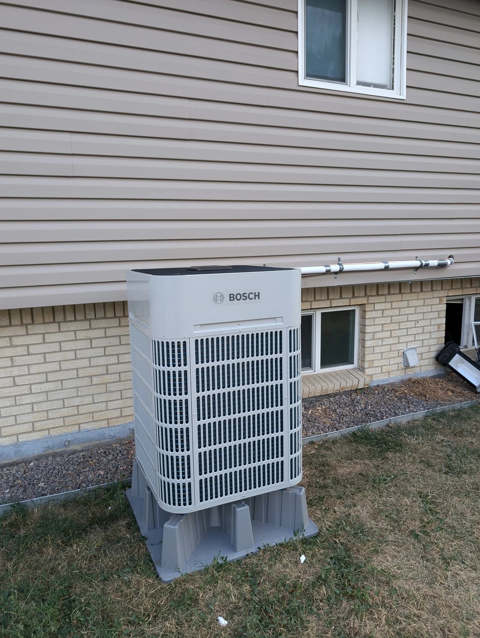 A Bosch outdoor unit on a composite pad on the lawn beside tan siding and a brick foundation