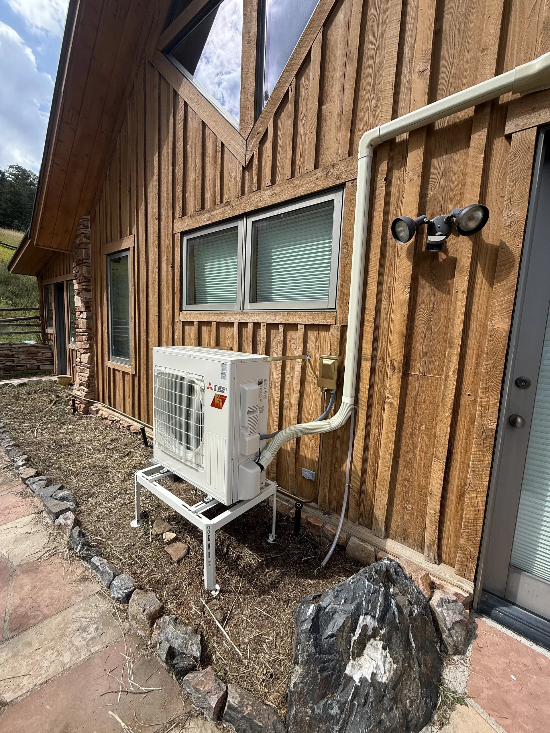 Mitsubishi compact outdoor unit on a stand beside a cedar board-and-batten wall, with a white line-set cover