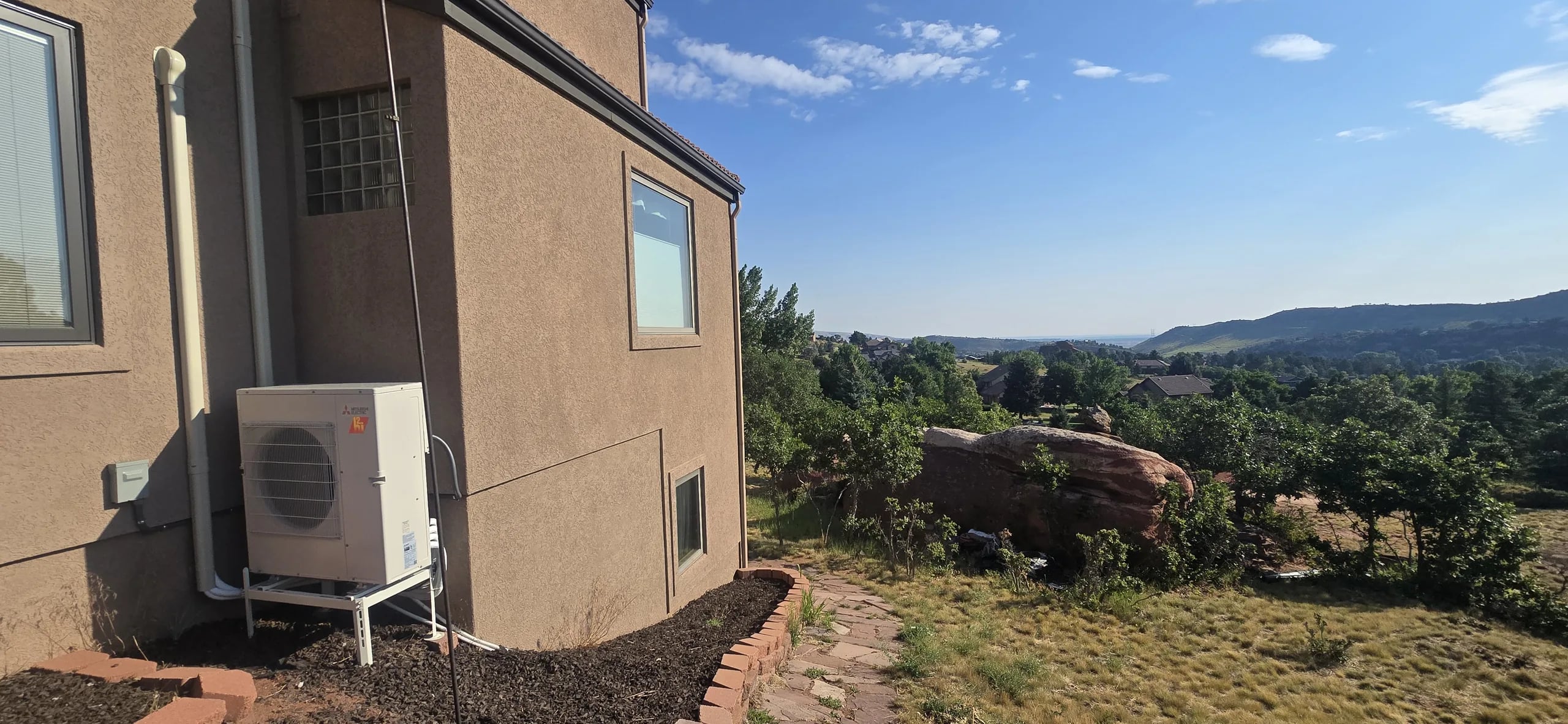 Mini-split outdoor unit on a stand beside a stucco house, looking out over pine-dotted foothills and red rocks