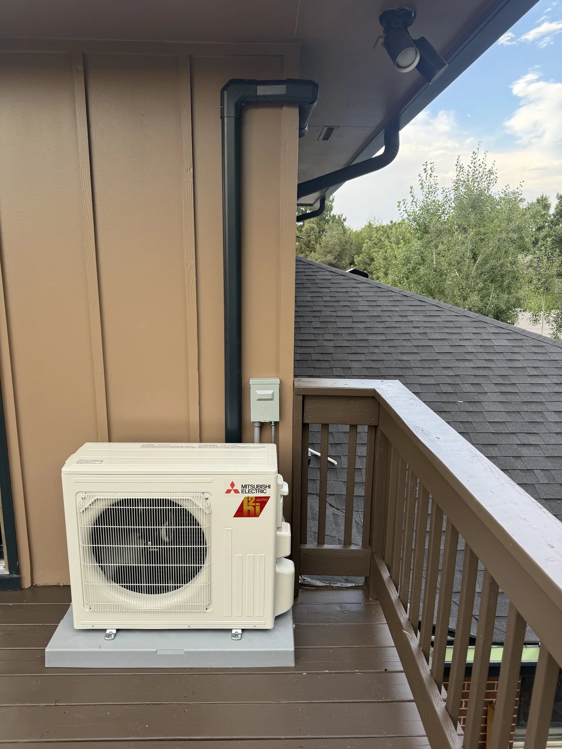 Mitsubishi outdoor unit on a pad on an upper wood deck with a brown railing and a downspout, trees beyond