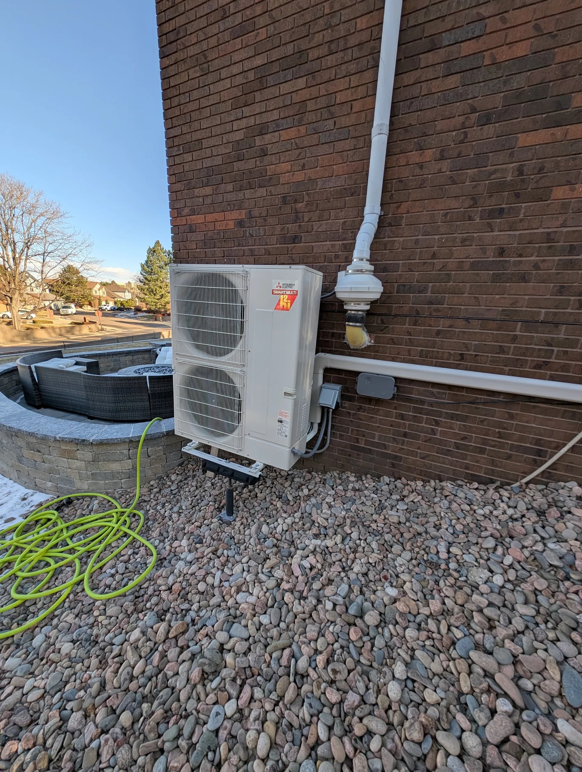 Mitsubishi two-fan outdoor unit beside a red brick wall in a river-rock bed, with a green garden hose