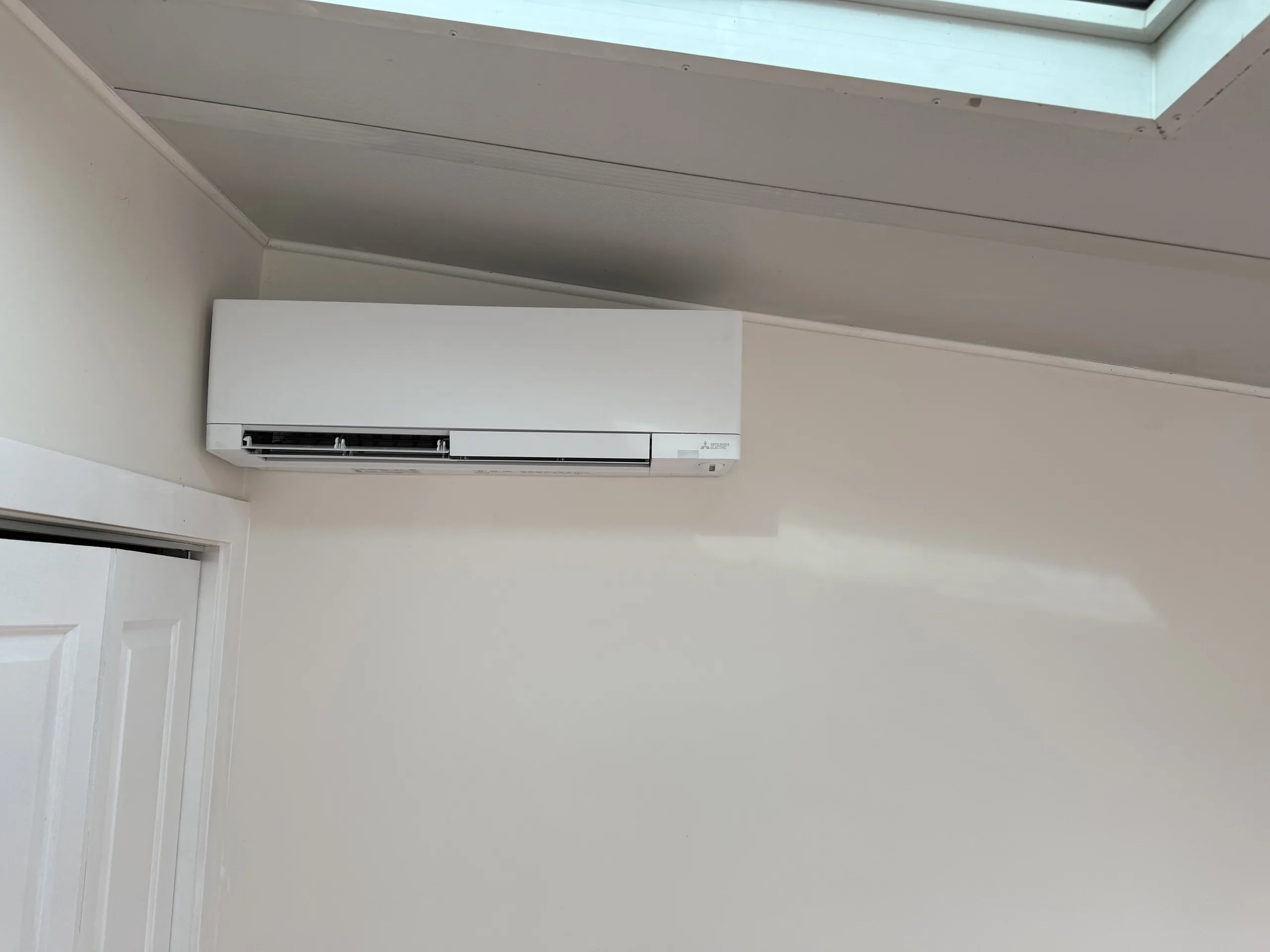 White Mitsubishi wall-mounted indoor unit on a white wall below a skylight, beside a closet door