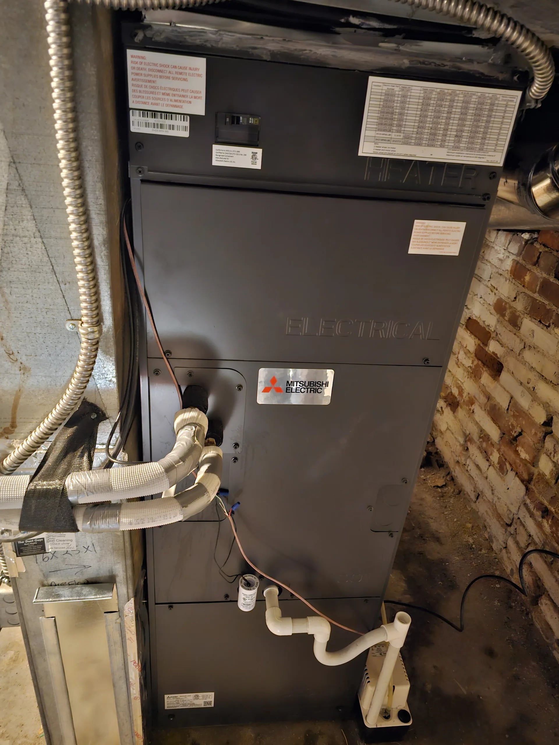 Black Mitsubishi air handler in a basement beside an exposed brick wall, with the refrigerant line and drain connected